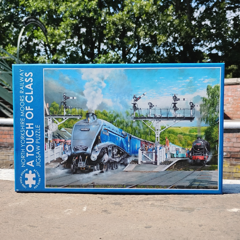 A boxed jigsaw puzzle titled 'A Touch of Class' by North Yorkshire Moors Railway, featuring a blue steam locomotive on a station platform. The box is photographed outdoors on a paved surface with trees and a brick wall in the background.