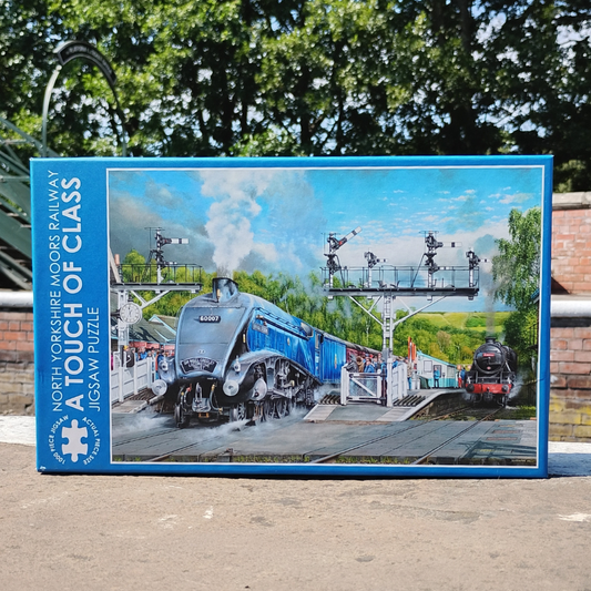 A boxed jigsaw puzzle titled 'A Touch of Class' by North Yorkshire Moors Railway, featuring a blue steam locomotive on a station platform. The box is photographed outdoors on a paved surface with trees and a brick wall in the background.
