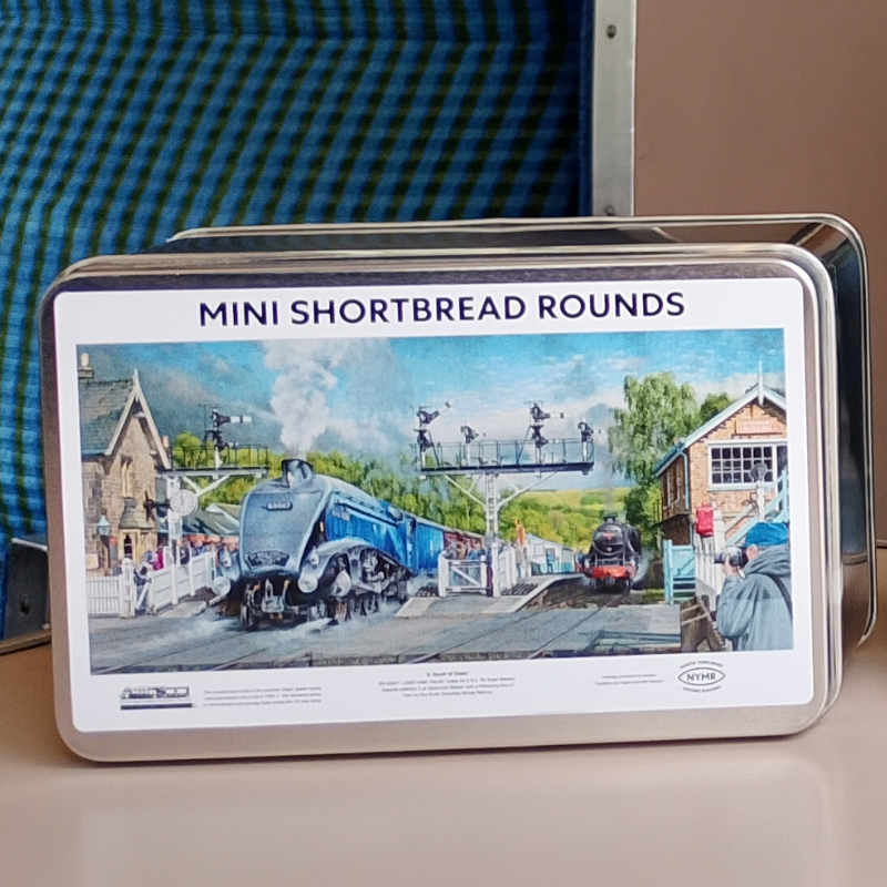 A rectangular metal tin labeled 'MINI SHORTBREAD ROUNDS' with a printed vintage train station scene on the front, photographed on a tabletop with a blue plaid fabric and beige wall in the background.
