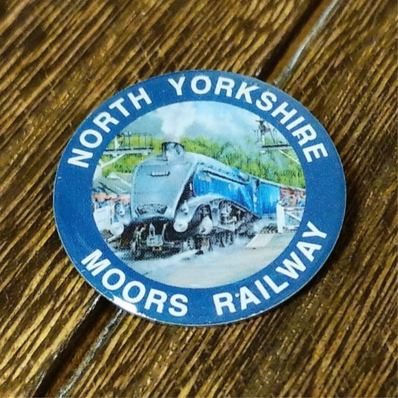 Circular pin badge on a wooden background. The pin badge has a painting illustration of Sir Nigel Gresley with a blue border and white text.