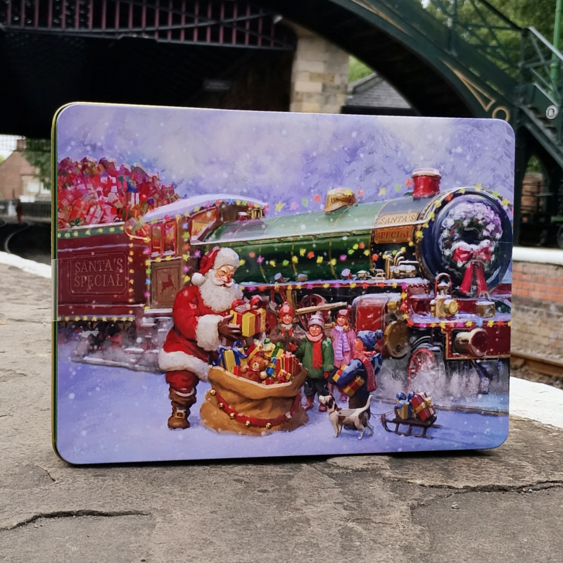 A square metal tin with rounded corners, featuring a colorful Christmas illustration of Santa Claus, children, a decorated steam train, and gifts in the snow. The tin is photographed standing on a stone railway platform.