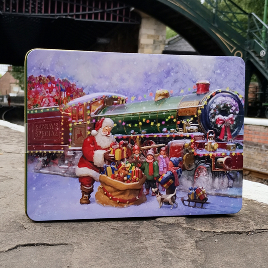 A square metal tin with rounded corners, featuring a colorful Christmas illustration of Santa Claus, children, a decorated steam train, and gifts in the snow. The tin is photographed standing on a stone railway platform.