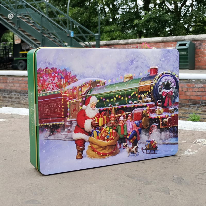 A rectangular Christmas-themed tin box with a colorful illustration of Santa Claus, children, and a festive train in the snow, photographed outdoors on a paved surface with a brick wall and greenery in the background.