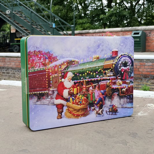 A rectangular Christmas-themed tin box with a colorful illustration of Santa Claus, children, and a festive train in the snow, photographed outdoors on a paved surface with a brick wall and greenery in the background.