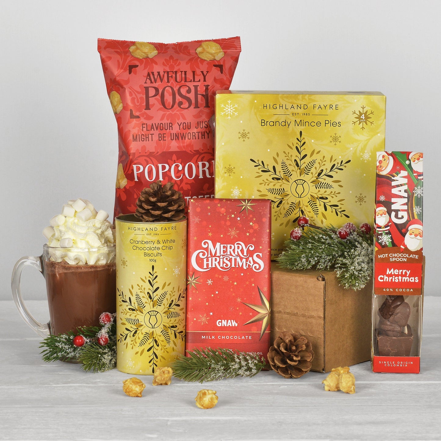A curated gift basket of treats and food items arranged together on a light gray surface against a white background. The set includes a red bag of Awfully Posh Popcorn, a yellow box of Highland Fare Brandy Mince Pies, a yellow can of Highland Fare Cranberry & White Chocolate biscuits, a red box of GNAW Merry Christmas Milk Chocolate, a box of GNAW Hot Chocolate, a glass mug filled with hot chocolate and whipped cream, and small holiday greenery and popcorn scattered around.