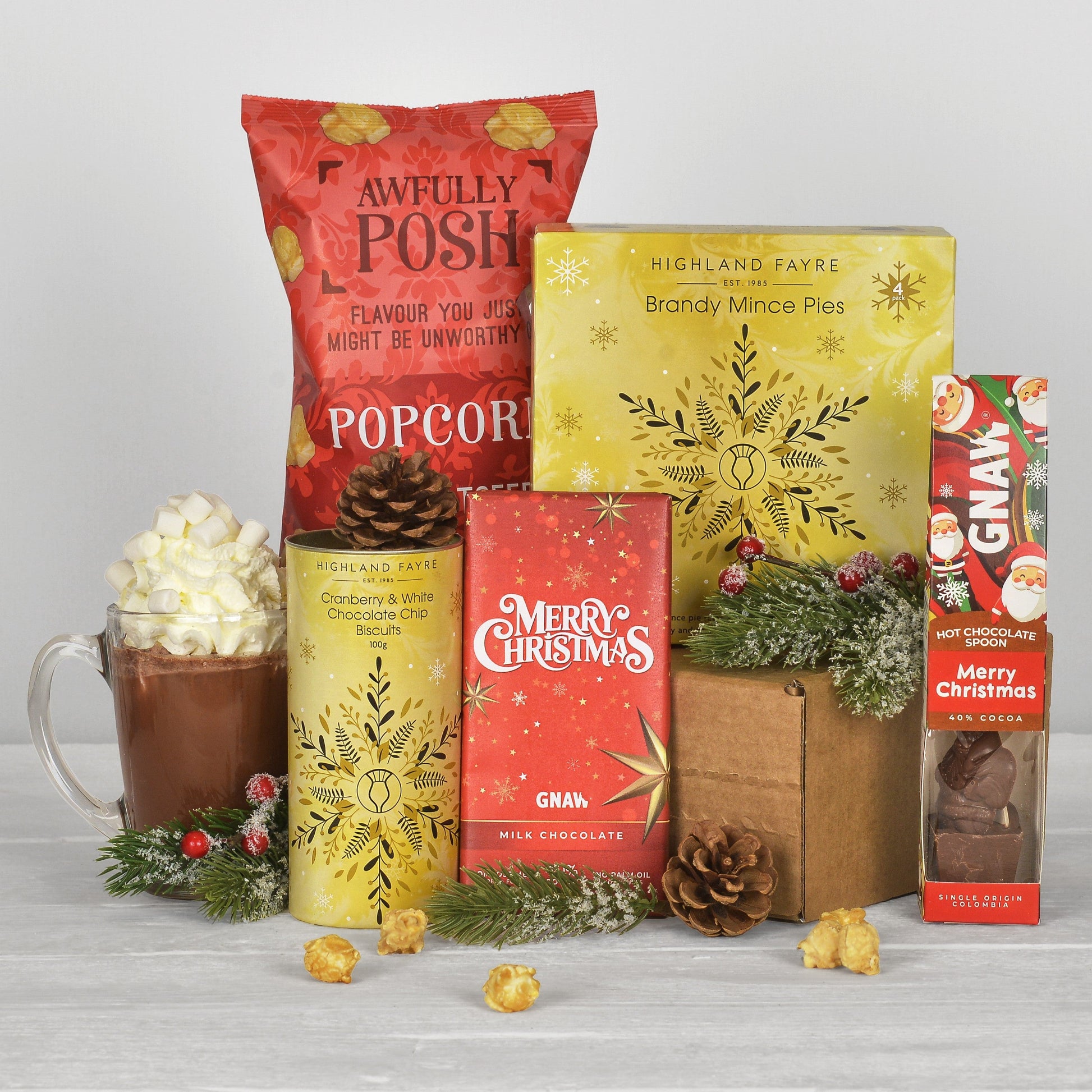 A curated gift basket of treats and food items arranged together on a light gray surface against a white background. The set includes a red bag of Awfully Posh Popcorn, a yellow box of Highland Fare Brandy Mince Pies, a yellow can of Highland Fare Cranberry & White Chocolate biscuits, a red box of GNAW Merry Christmas Milk Chocolate, a box of GNAW Hot Chocolate, a glass mug filled with hot chocolate and whipped cream, and small holiday greenery and popcorn scattered around.