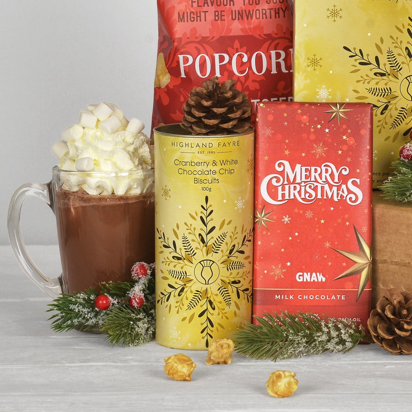 A festive gift assortment featuring Highland Fayre Cranberry & White Chocolate Chip Biscuits in a yellow tin, GNAW Milk Chocolate in a red box, popcorn, and a mug of hot chocolate topped with marshmallows, styled with pine branches and pinecones on a light gray surface.
