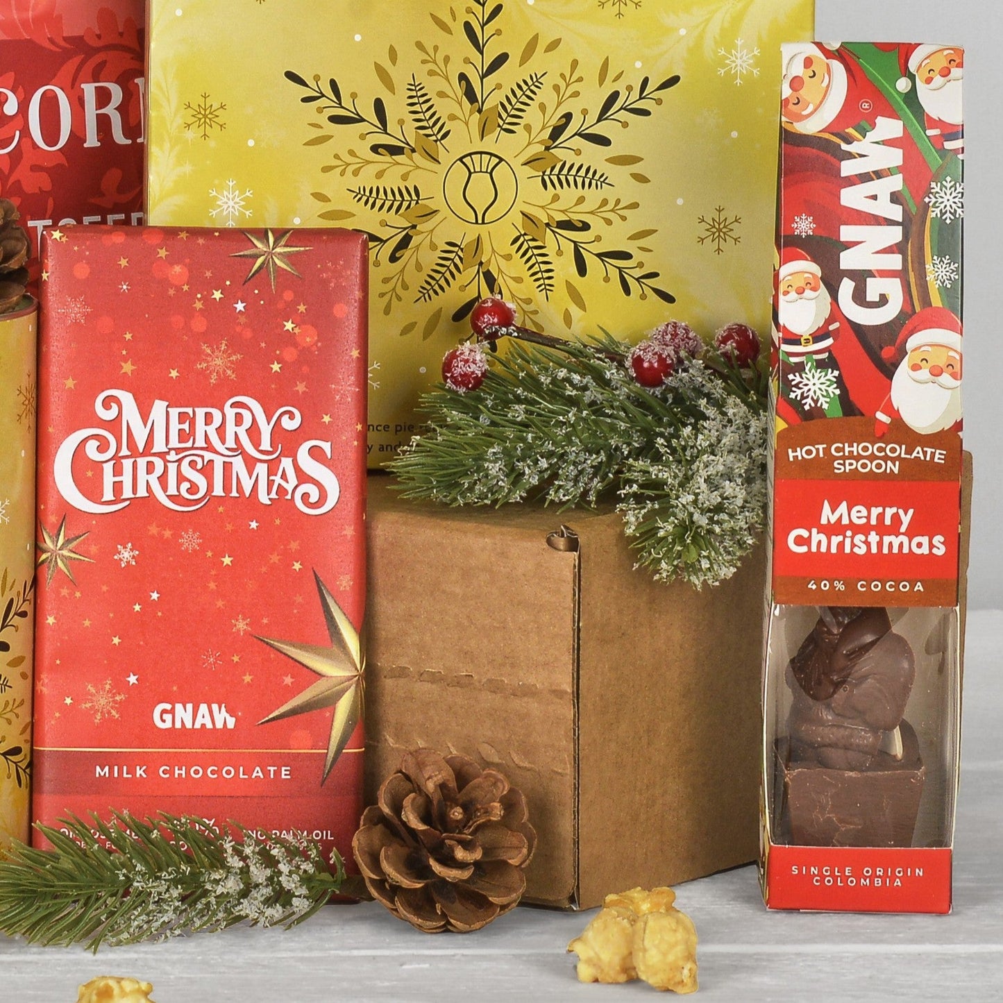 A Christmas-themed chocolate gift set featuring a red box of GNAW Merry Christmas milk chocolate and a GNAW Merry Christmas hot chocolate spoon, styled with green pine branches, a pinecone, and a brown box on a light gray surface.