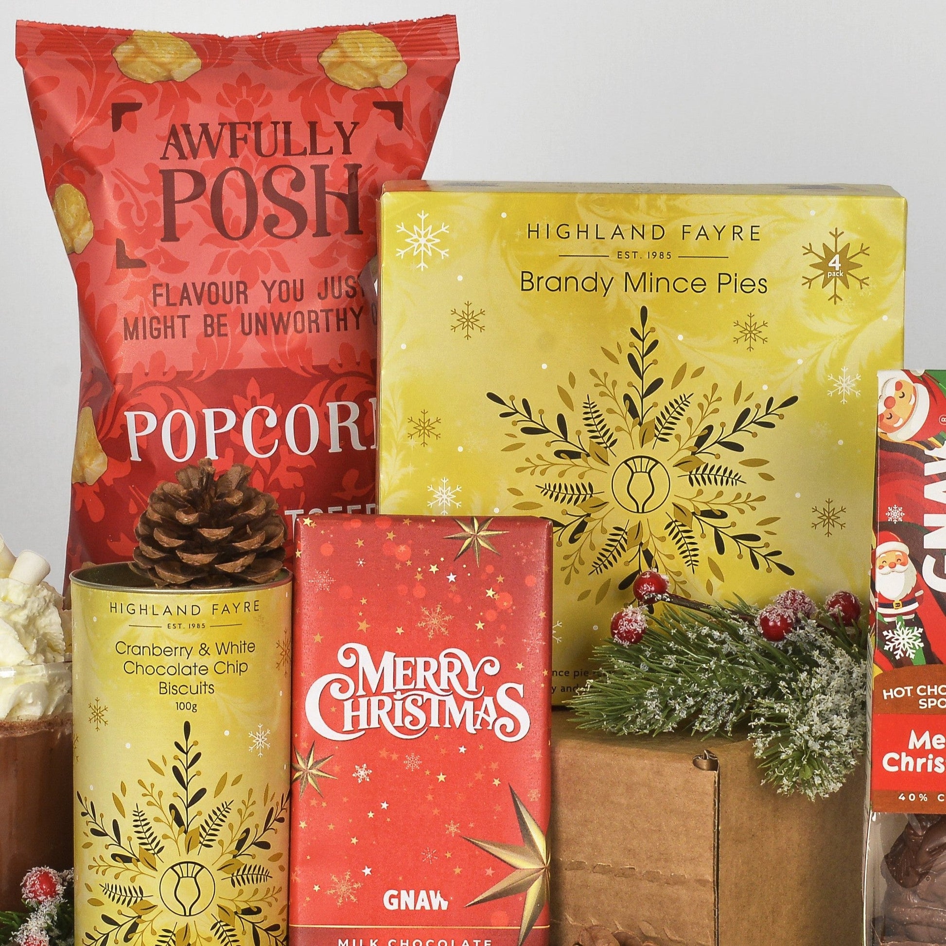 A Christmas-themed gift package featuring Highland Fayre products including a large yellow box of Brandy Mince Pies, a red bag of Awfully Posh Popcorn, a red box of Merry Christmas Gnaw milk chocolate, and a can of cranberry and white chocolate chip biscuits, styled with a pinecone and greenery on a neutral background.