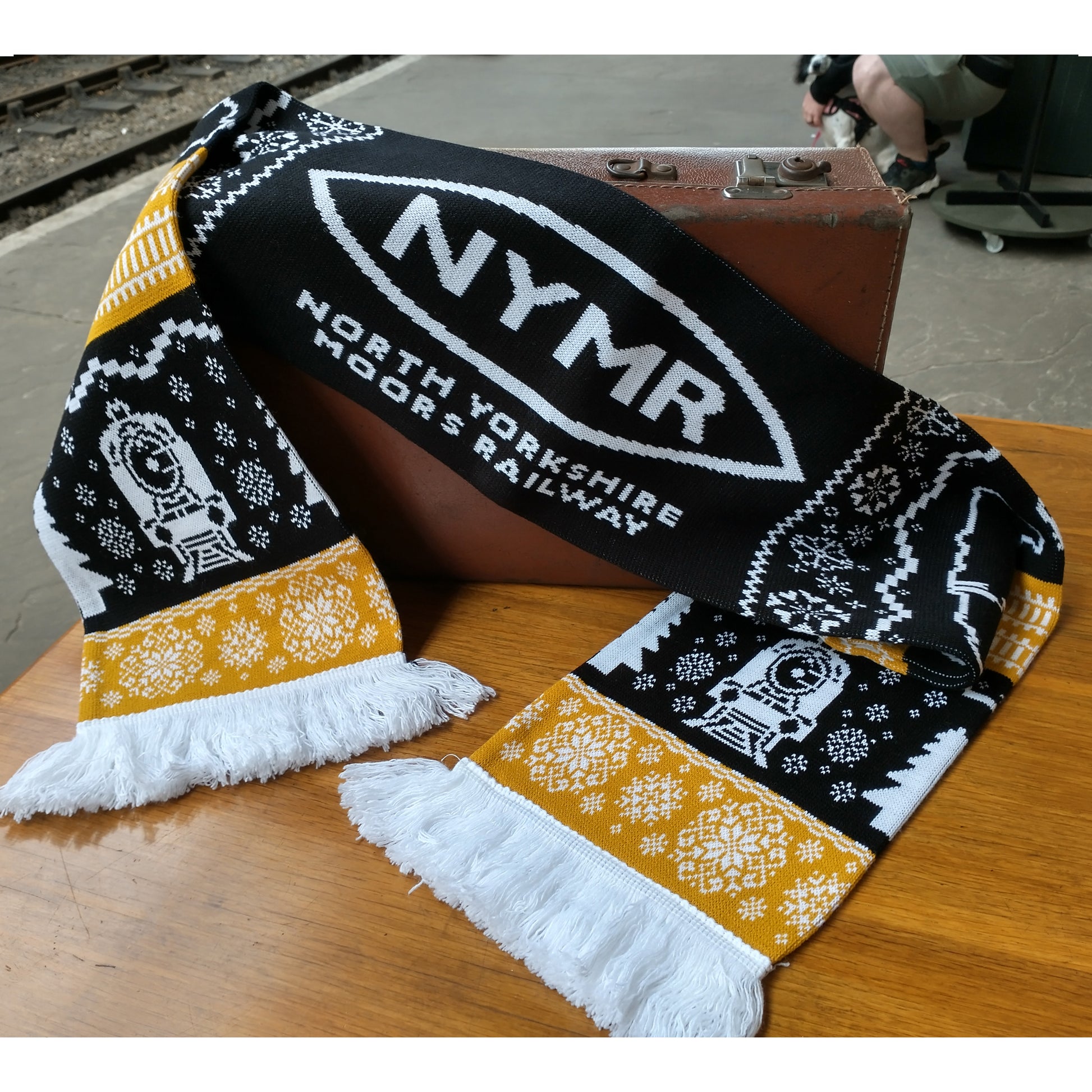 A knitted scarf in black, white and golden threads. With the NYMR logo in white on black, and loco details. Snowflakes and track motifs are white on a golden background.