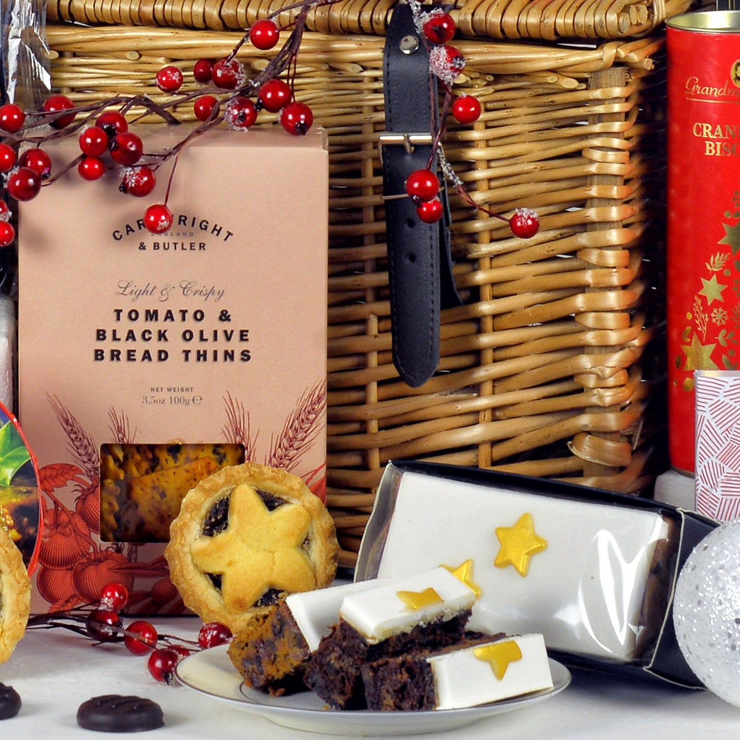 Assorted food items including Tomato and Olive bread thins, a mince pie, and slices of Christmas cake in front of a wicker basket.