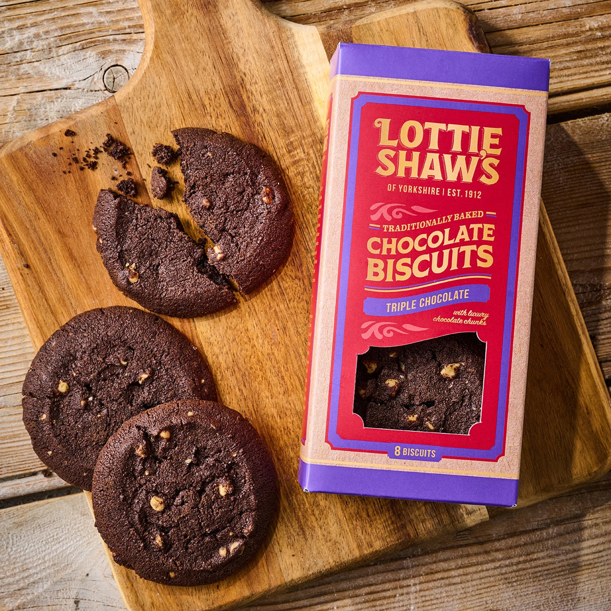 A Box of biscuits aside three biscuits out of the box on a wooden board, one of them is broken and there are tasty looking crumbs to the side.....