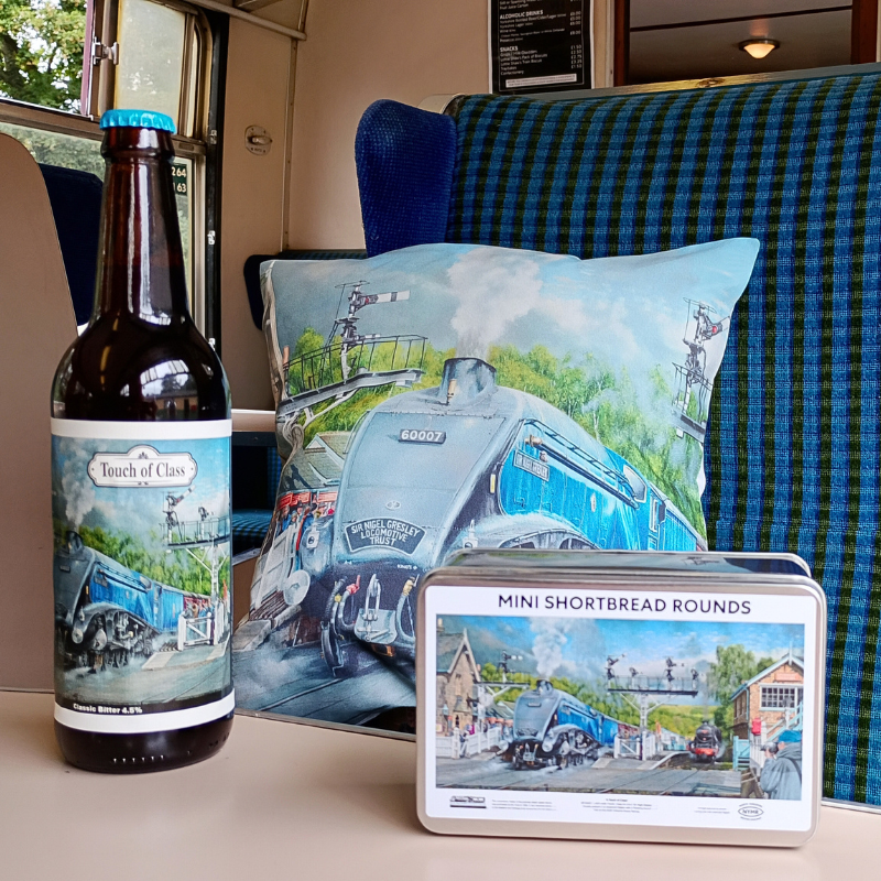 A dark brown bottle of Touch of Class beer, a decorative cushion, and a metal tin of Mini Shortbread Rounds, all featuring matching vintage railway illustrations, arranged on a train seat next to a blue plaid cushion.