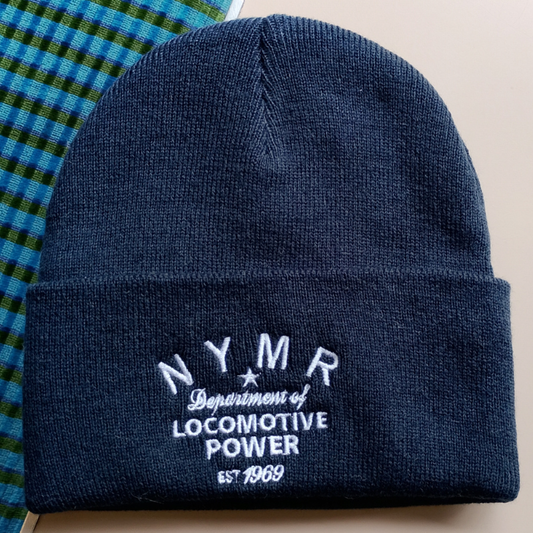 A navy blue knit beanie with white embroidered text reading 'NYMR Department of Locomotive Power Est. 1969', shown folded on a light beige surface with a blue-green plaid fabric visible at the upper left.