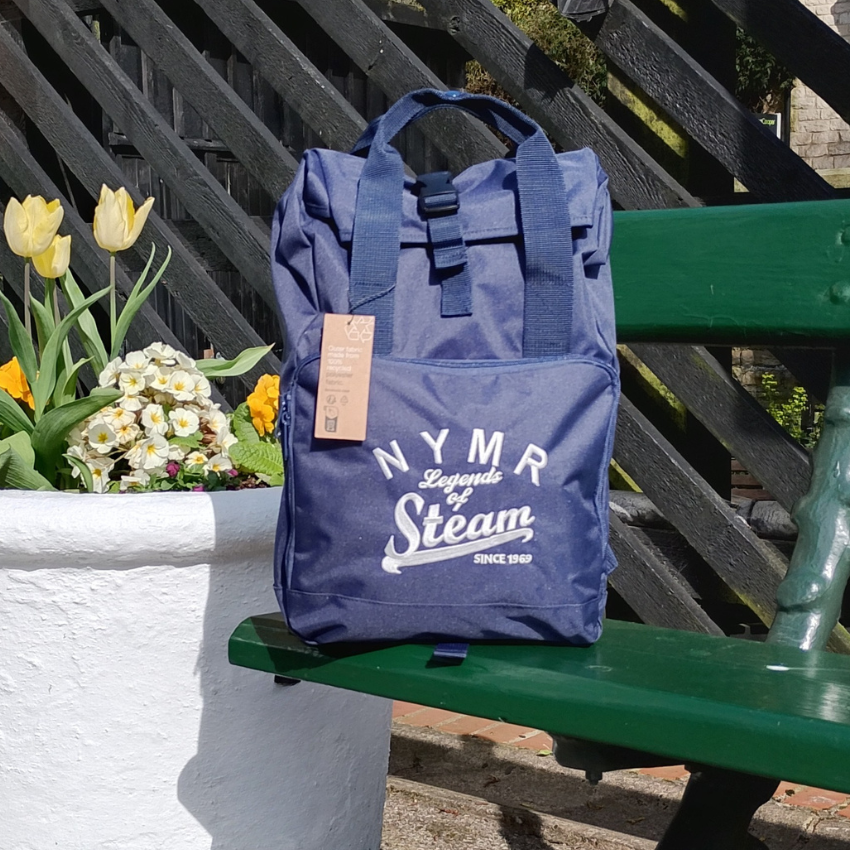 Blue backpack with 'NYMR Legends of Steam' text on a green bench with flowers in the background