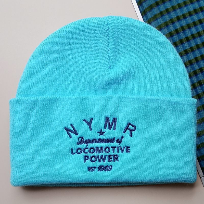 A bright turquoise knit beanie hat with dark blue embroidered text reading 'NYMR Department of Locomotive Power est. 1969' on the front cuff, shown on a light beige surface with a blue-and-green plaid fabric partially visible at the top right.