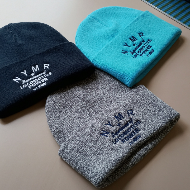 Three knit beanie hats in navy, turquoise, and heather gray, each embroidered with 'NYMP National Yacht Club Locomotive Power 1968' on the cuff, arranged on a beige surface.