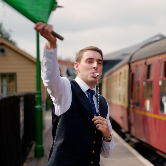 A train guard blowing a whistle and waving a green flag on the North Yorkshire Moors Railway.