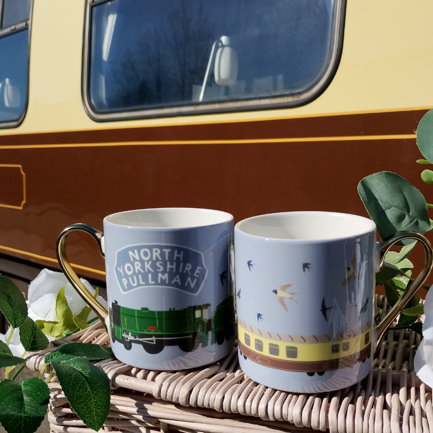 Two mugs with train designs on a woven surface in front of a vintage-style vehicle.