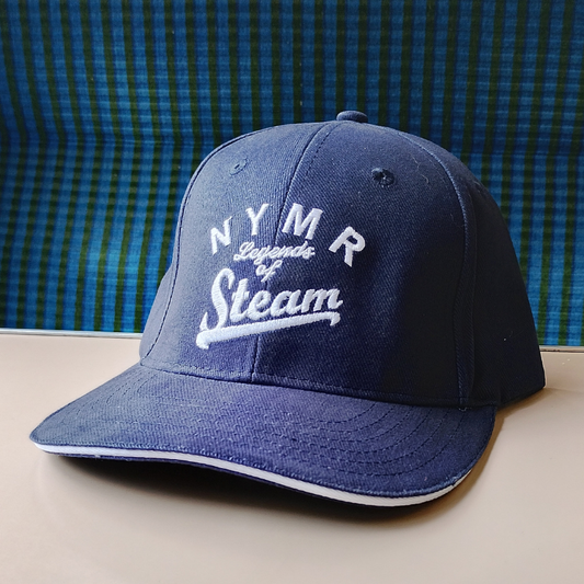 A navy blue baseball cap with white embroidered text reading 'NYMR Legends of Steam' on the front, resting on a light beige surface with a blue-green plaid background.