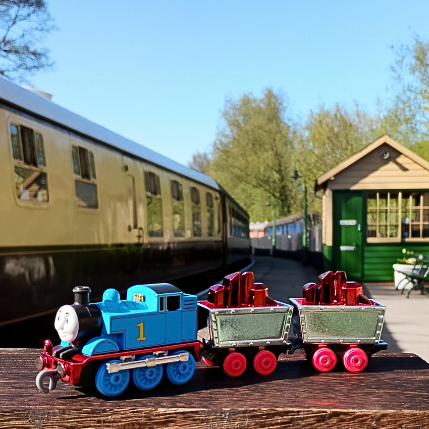Model train set with colorful engine and cars on a wooden platform, with a train station in the background.