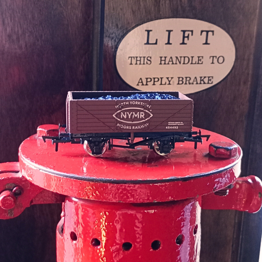 Model train car with 'NYMR' logo on a red platform, with a wooden background and 'LIFT THIS HANDLE TO APPLY BRAKE' sign.