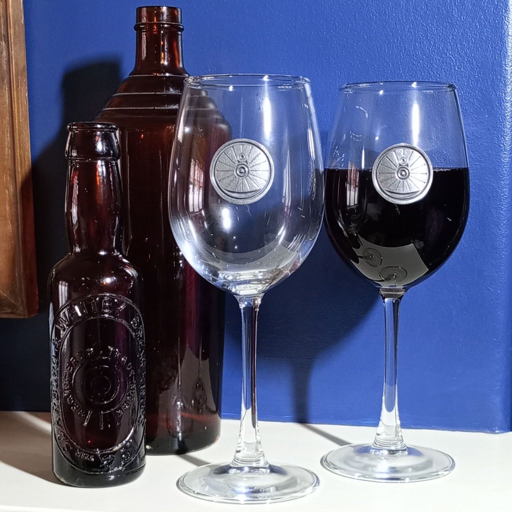 A pair of wine glasses with a pewter wheel on the front, one glass is either half full or half empty of red wine. 