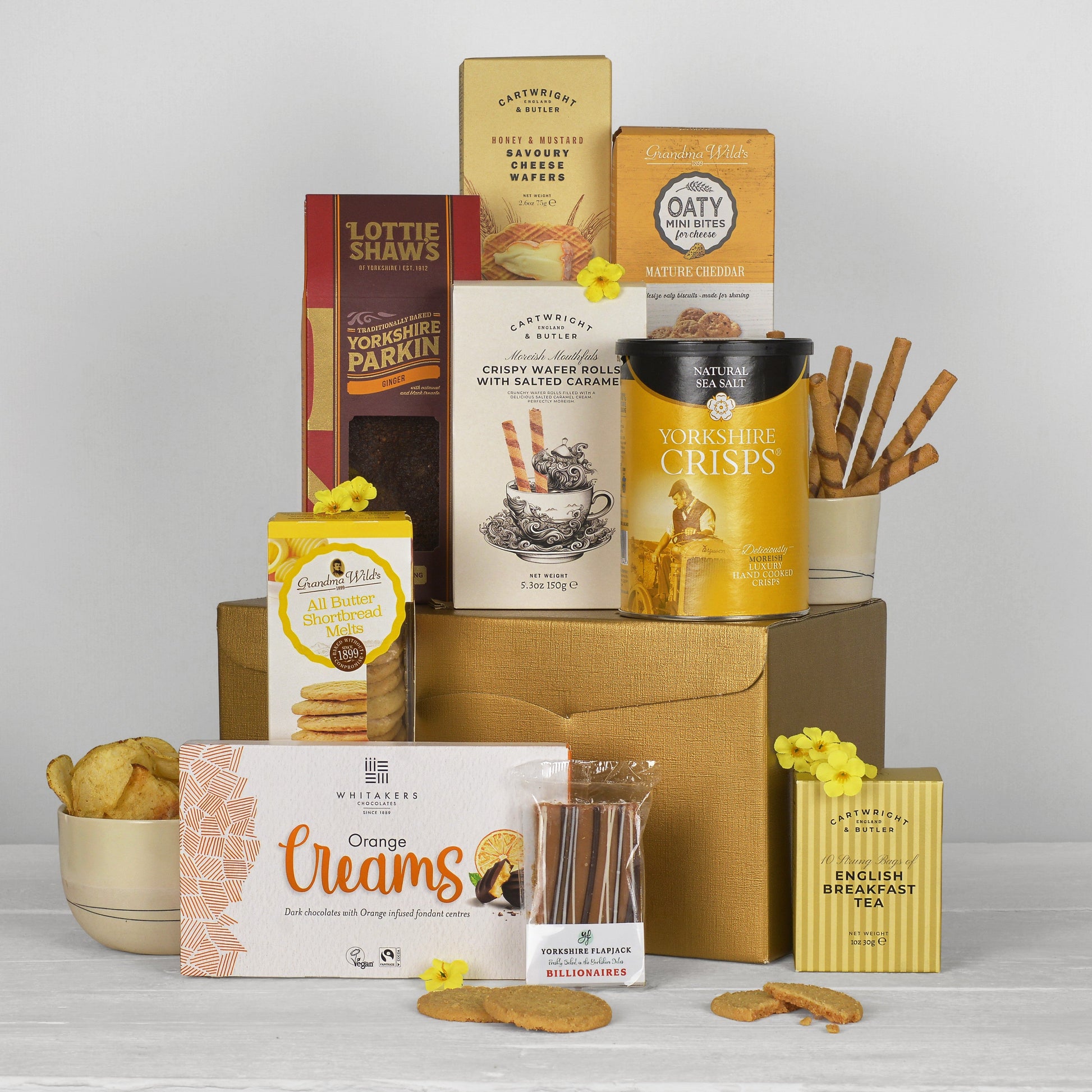 A curated gift hamper of assorted Yorkshire snacks and biscuits arranged on a golden cardboard box against a light gray studio background. Visible products include Lottie Shaws bakes, Yorkshire Crisps, Oatmeal Crispin, English Breakfast Toffee, and others, with biscuits and sticks in bowls and packaging.