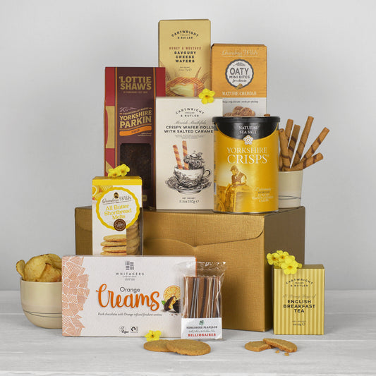 A curated gift hamper of assorted Yorkshire snacks and biscuits arranged on a golden cardboard box against a light gray studio background. Visible products include Lottie Shaws bakes, Yorkshire Crisps, Oatmeal Crispin, English Breakfast Toffee, and others, with biscuits and sticks in bowls and packaging.