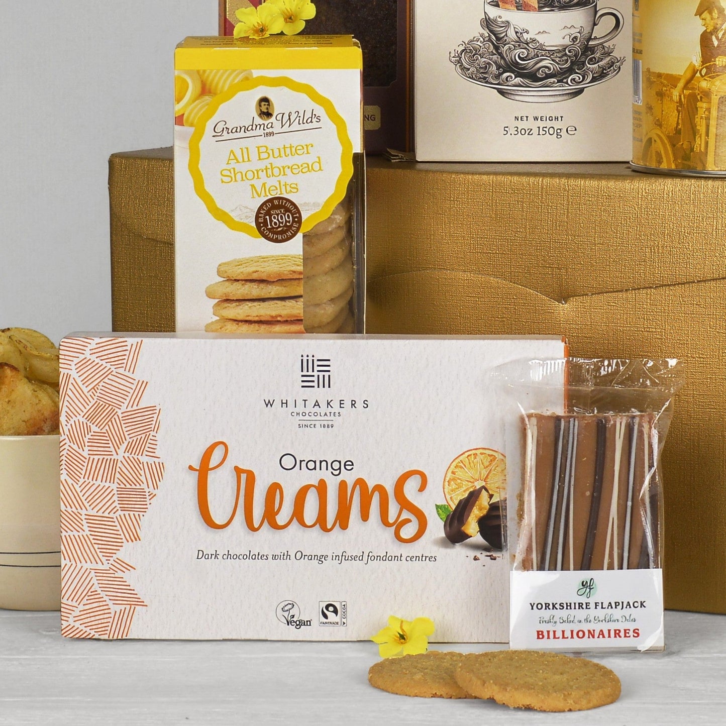A gift basket arrangement of baked goods and chocolates, featuring a large box of Whitakers Orange Creams in the foreground, a box of Grandma Wild's All Butter Shortbread Melt biscuits behind it, and a Billionaires Yorkshire Flapjack in front, set against a gold gift box on a white surface.