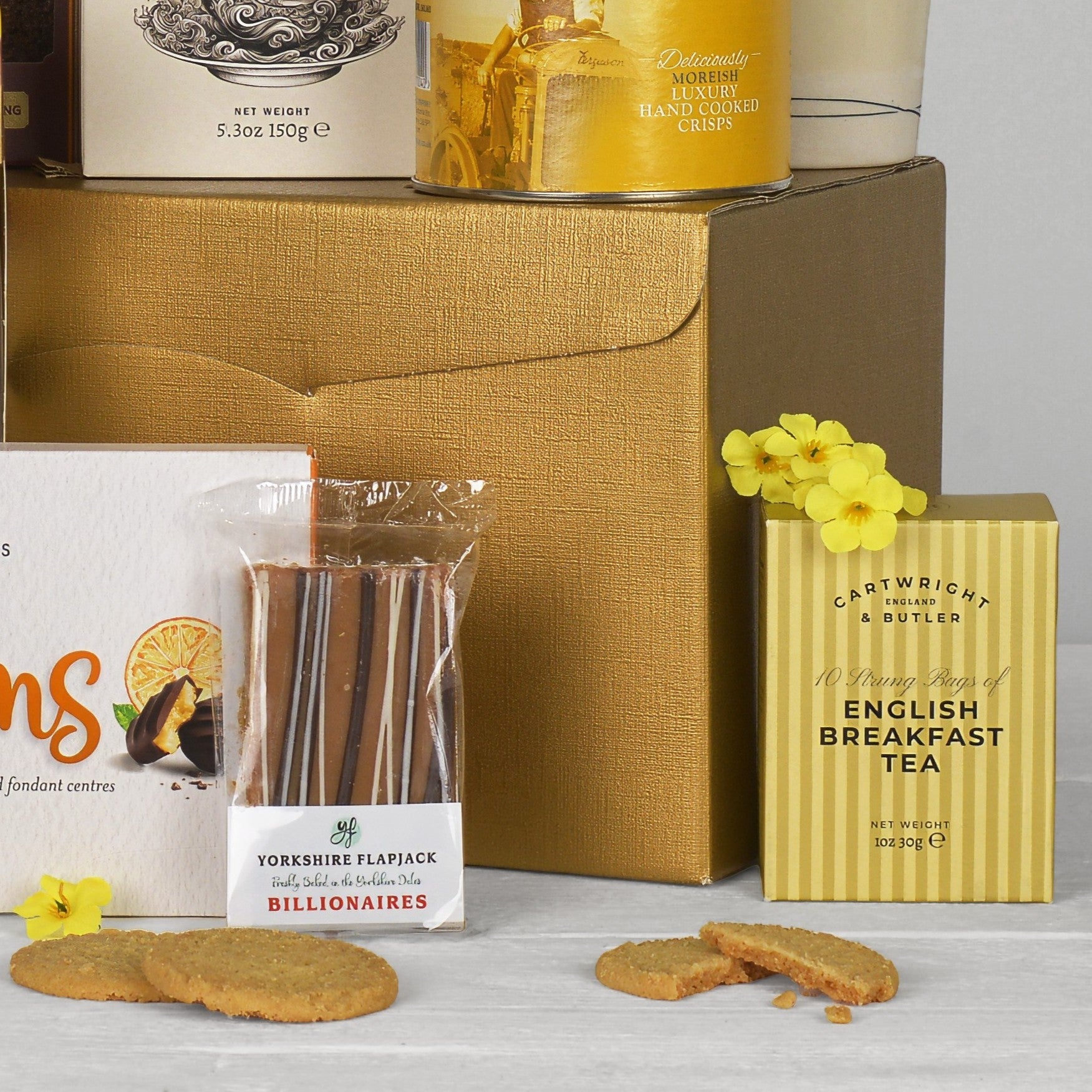A gift hamper arrangement featuring a large gold box, a box of Cartwright & Butler English Breakfast Tea, a package of Yorkshire Flapjack biscuits, and other assorted treats on a white surface with a light gray background.