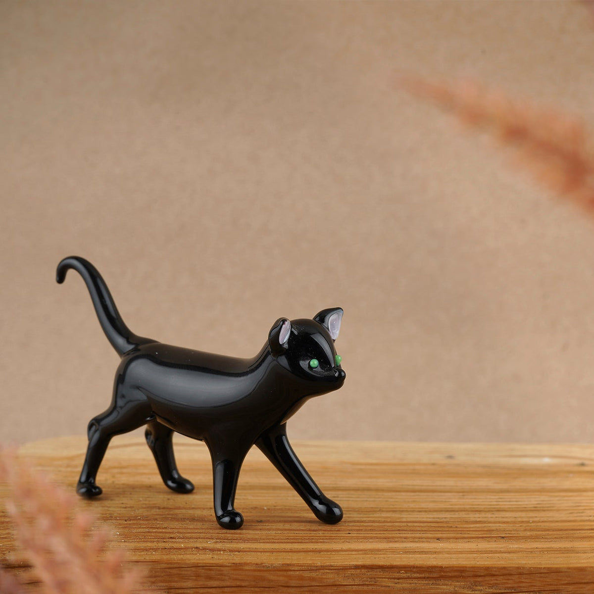A glass ornament of a black cat with tail high and curved and green eyes, on a wooden counter top.