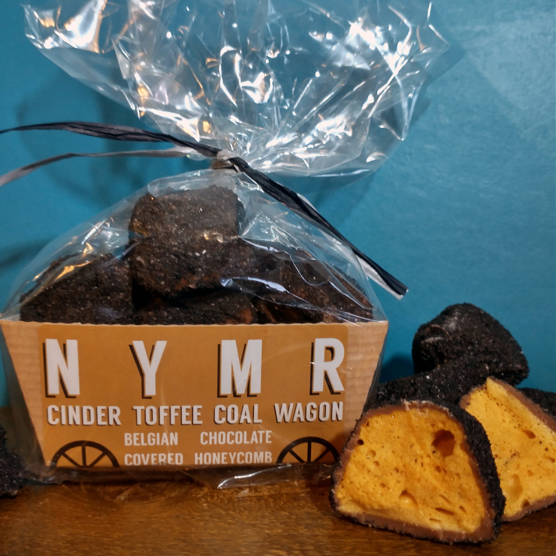 A cellophane bag containing lumps of chocolate coal in a brown railway style wagon. There are chunks of the coal next to the packet which have been cut open to show the golden honeycomb and thick chocolate coating. Black sherbet covers each piece.