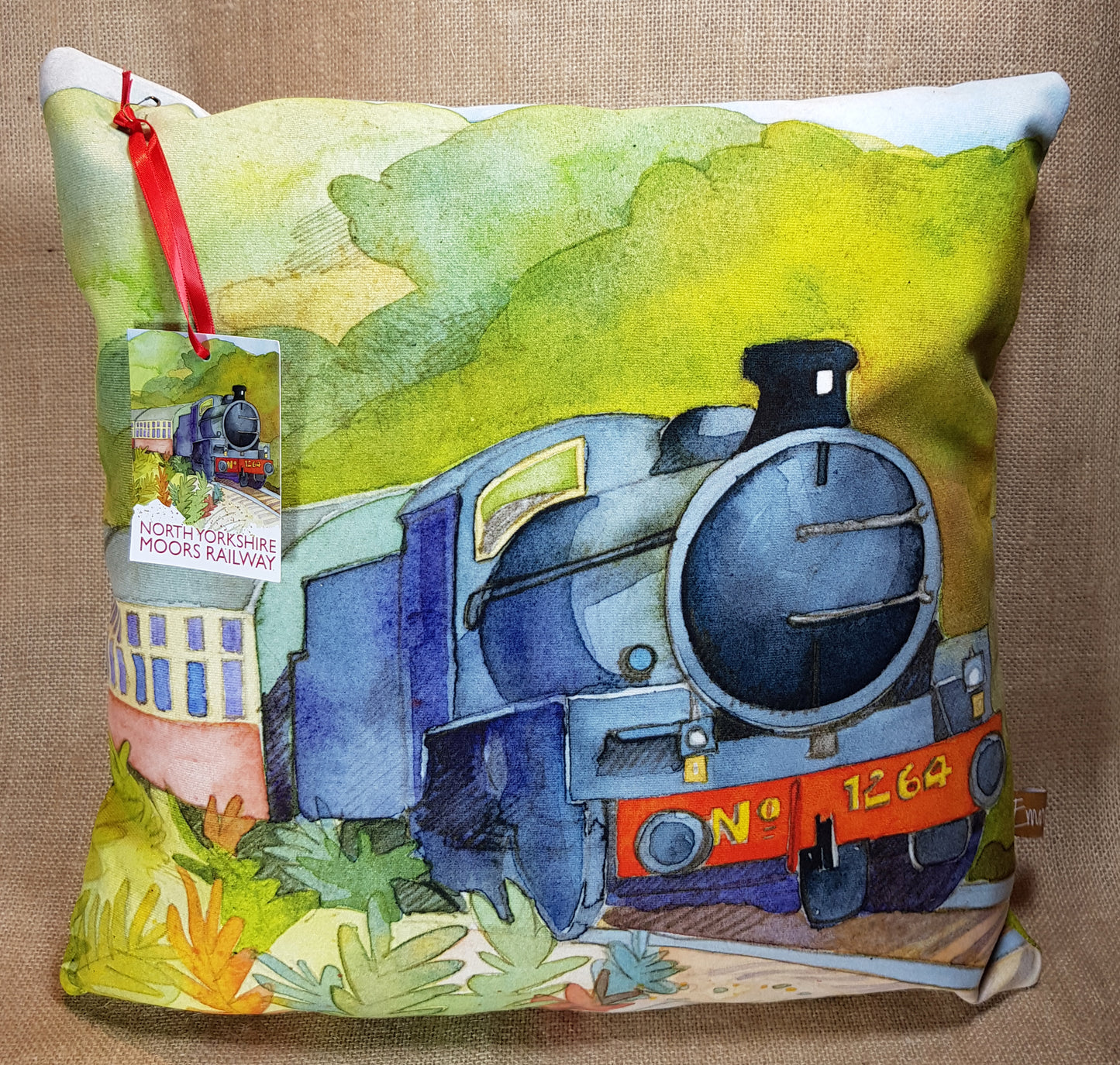 Square cushion with painting of steam train travelling through countryside and North Yorkshire Moors Railway label attached with red ribbon.