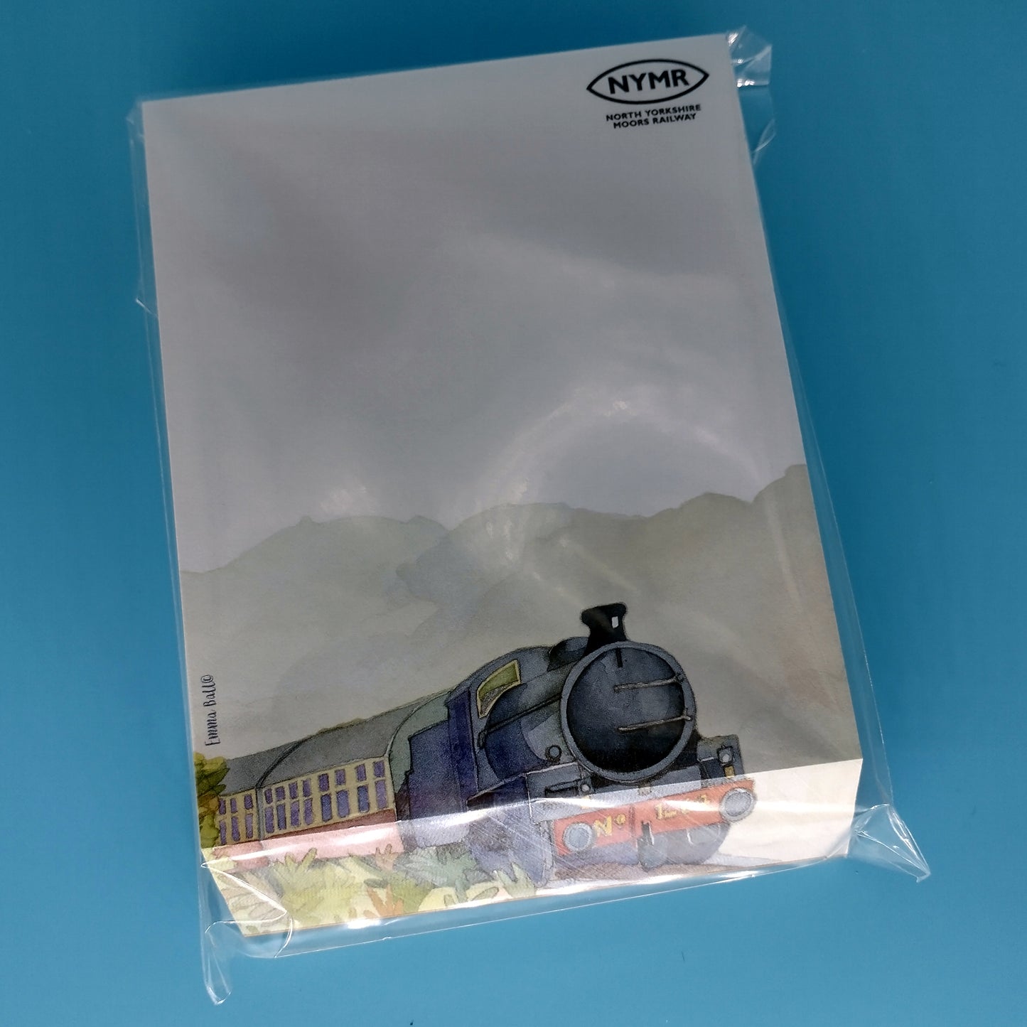 Note pad with painting of steam train travelling through countryside across bottom and N Y M R logo printed in black in top right hand corner.