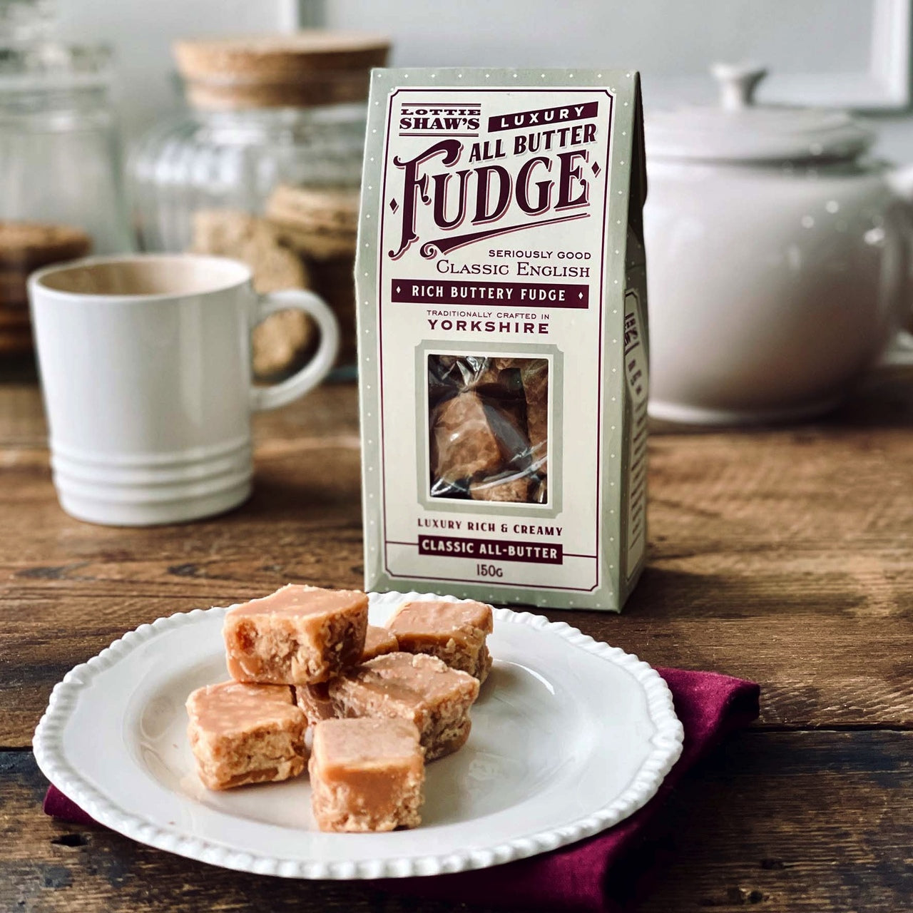 Packet of Lottie Shaw's luxury all butter fudge with contents showing through cellophane window. There is some fudge pictured on a plate on a kitchen table.