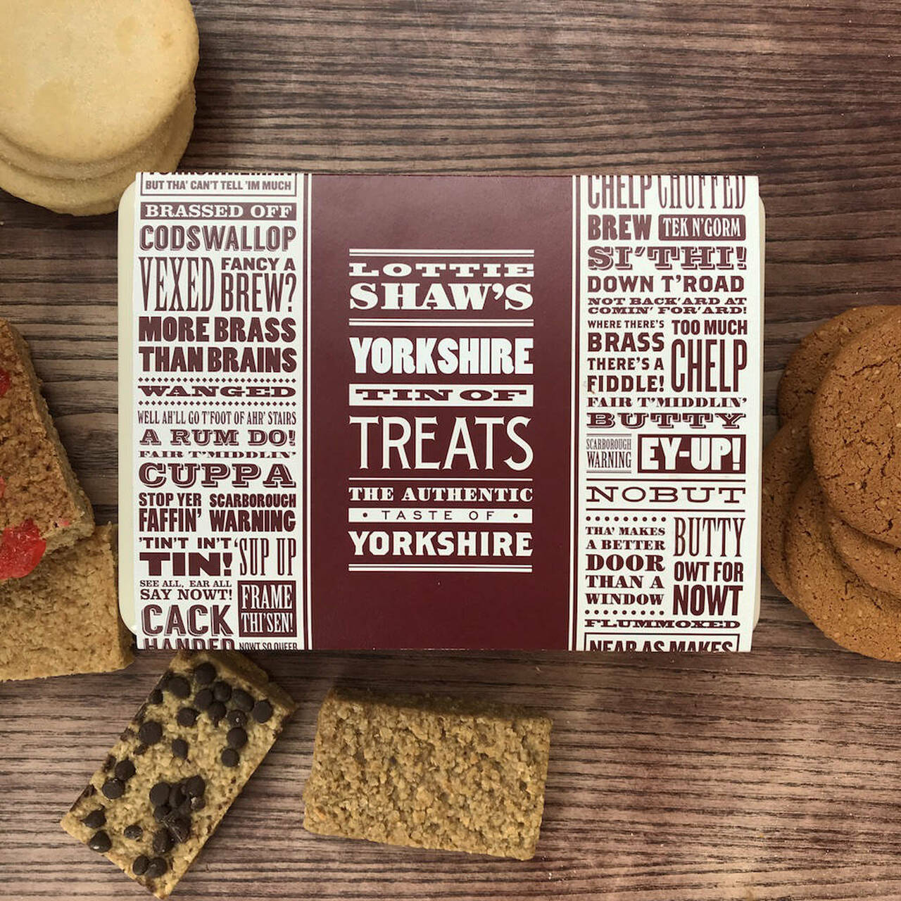 Lottie Shaw's oblong tin of biscuits and flapjacks with card sleeve printed with Yorkshire sayings and a selection of the contents shown on table.