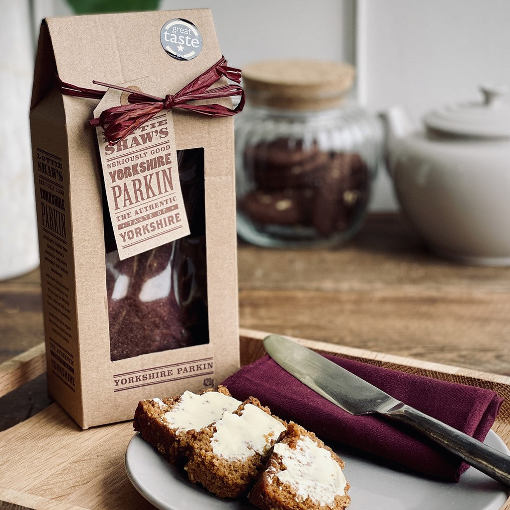 Packet of Lottie Shaw's seriously good Yorkshire parkin with contents showing through cellophane window. There is some buttered parkin pictured on a plate on a kitchen table.