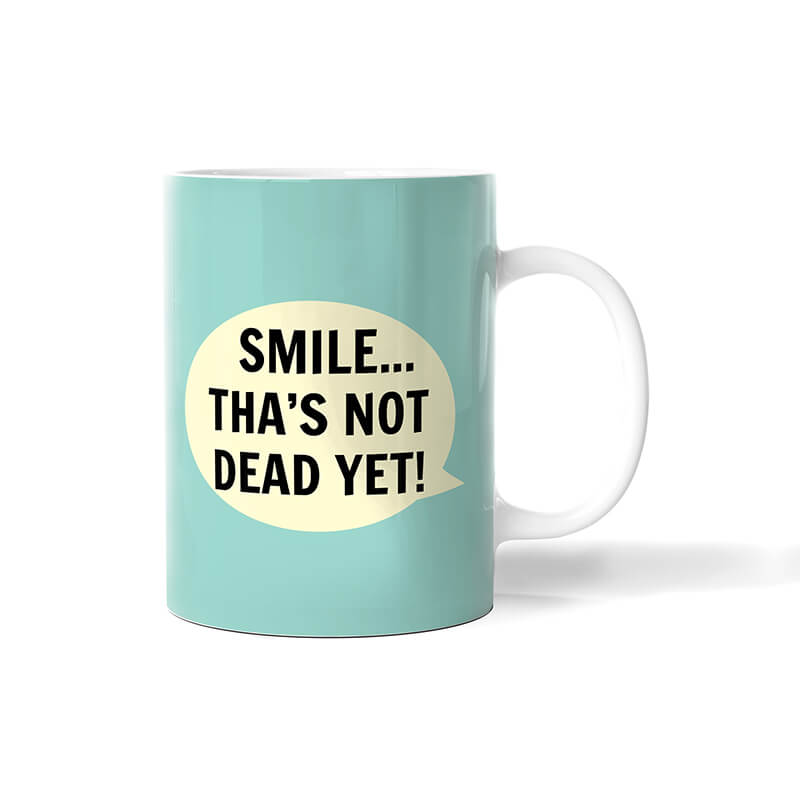Turquoise mug with white inside and handle and 'Smile ... tha's not dead yet!' printed in black in white speech bubble on side.